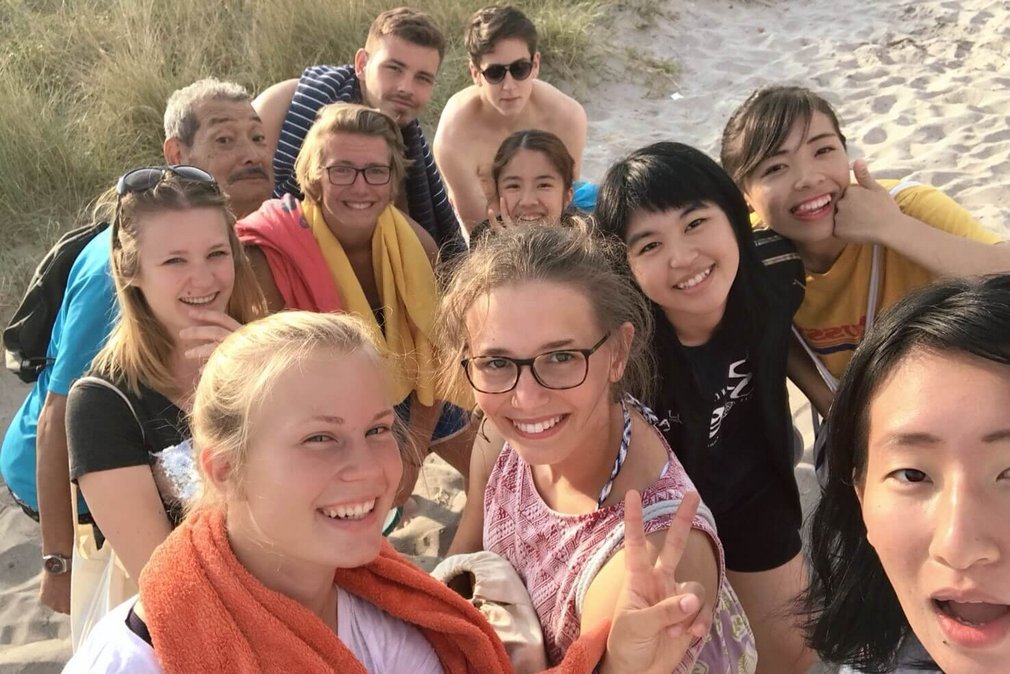 Eine Gruppe von Menschen posiert fuer ein Selfie am Strand und laechelt in die Kamera.