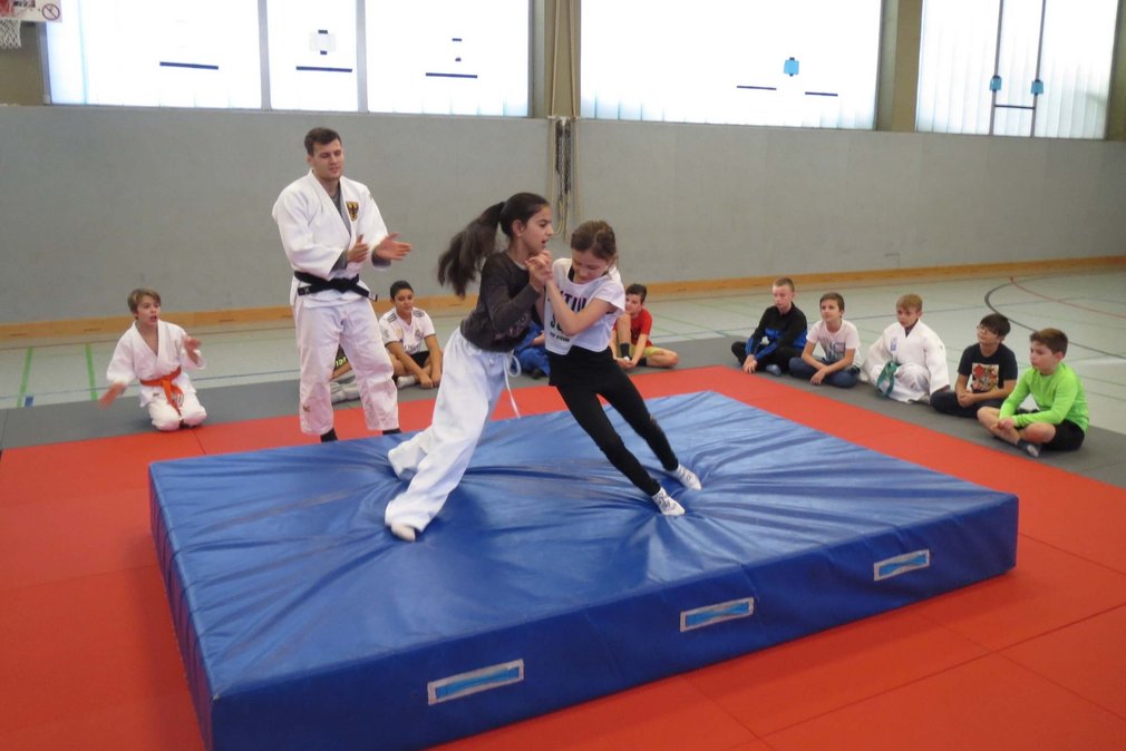 Zwei Maedchen bei einem Trainingskampf beim Tag des Judo. Andere Teilnehmenden sitzen auf dem Boden um sie herum.