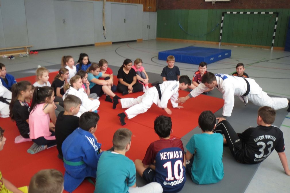 Ein Trainer und ein Schueler bei einem Trainingsspiel in Liegestuetzposition. Alle anderen sitzen um sie herum und schauen ihnen zu.