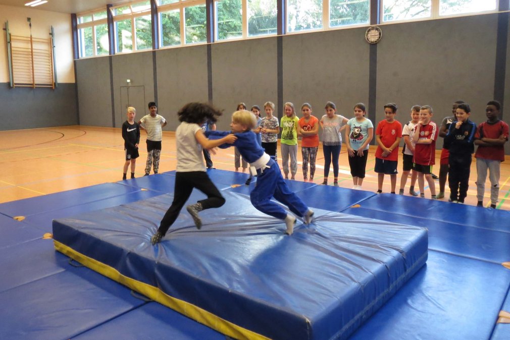 Zwei Jungs bei einem Trainingskampf beim Tag des Judo. Andere Teilnehmenden stehen drum herum und schauen ihnen zu.