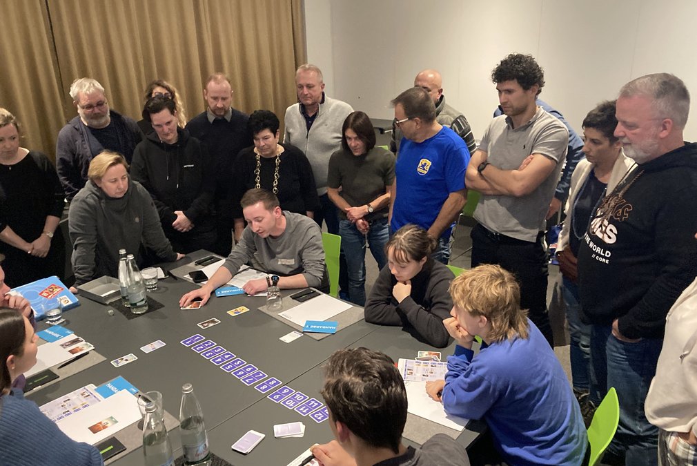 Viele Menschen sitzen und stehen um einen Tisch und spielen das Planspiel: Schau hin und sag was!
