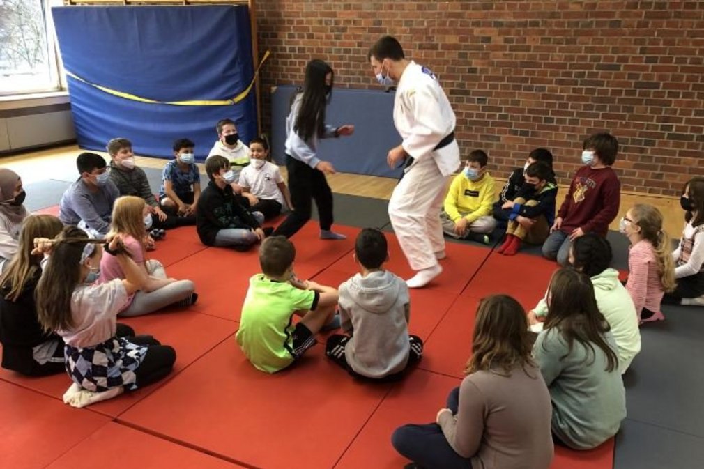 Ein Judotrainer und seine Schuelerin trainieren miteinander. Weitere Kinder sitzen im Kreis um sie herum.