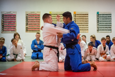 Zwei Judoka kaempfen gegeneinander. Weitere sitzen in einem Kreis um sie herum.
